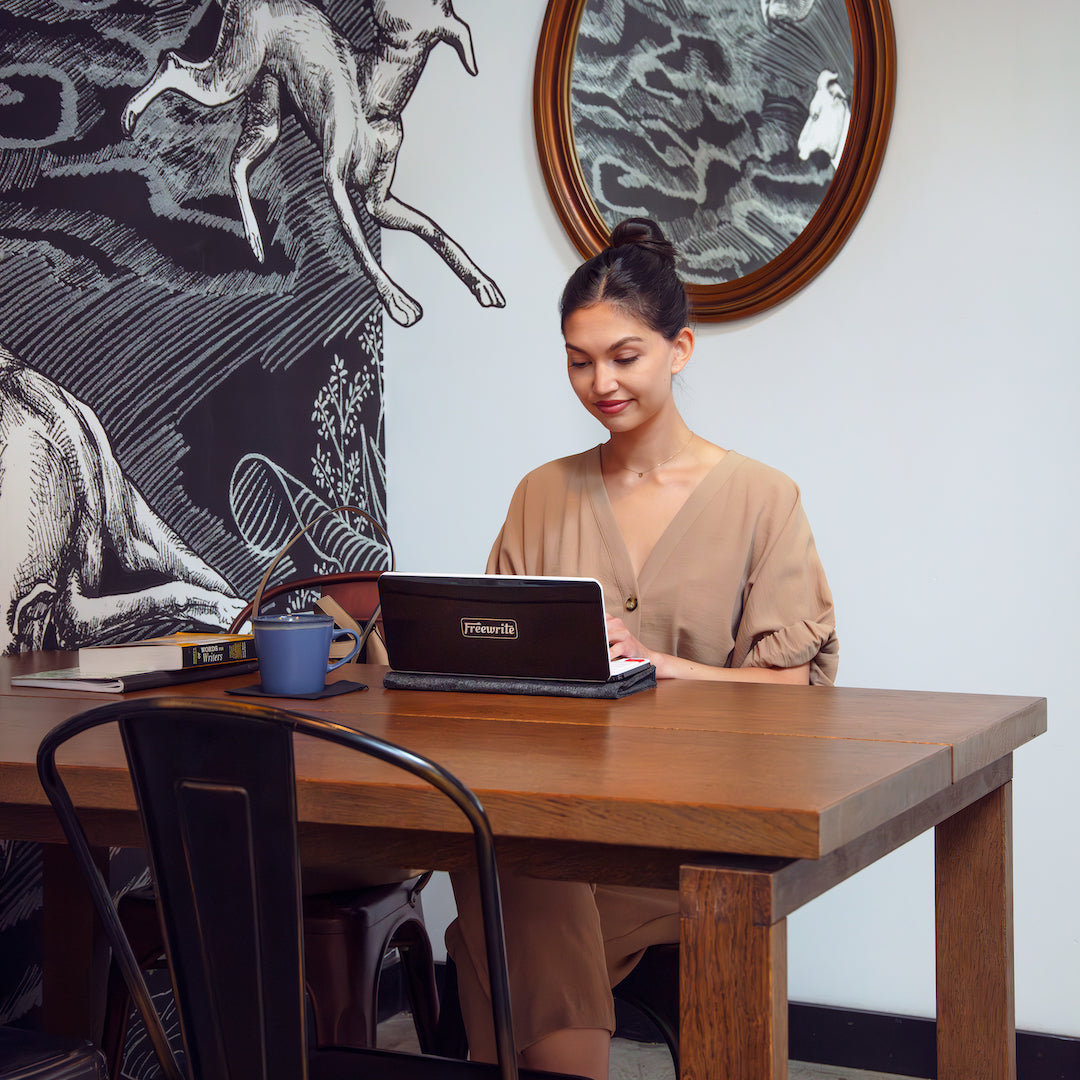 Woman typing on a laptop at a wooden table outdoors, beside a water bottle and books.