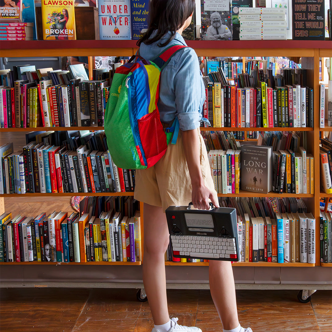 Person with colorful backpack and typewriter in front of a bookshelf in a bookstore.