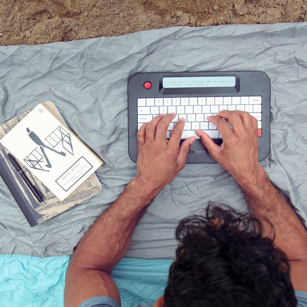 Person typing on a portable electronic typewriter with a notebook and pen beside them on a gray background.