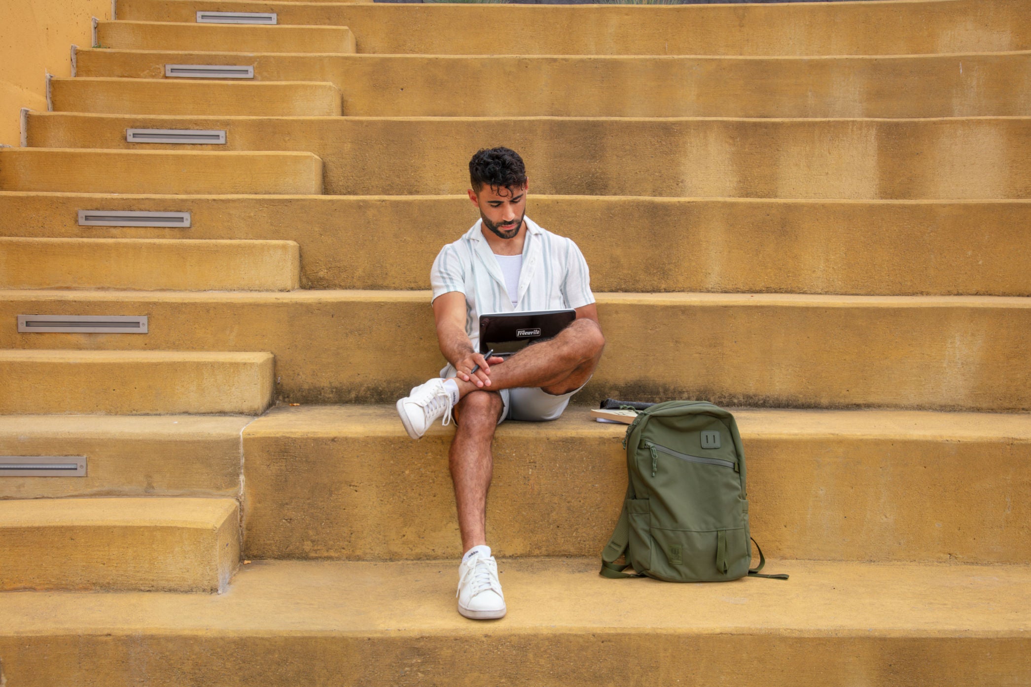 Man sitting on outdoor steps working on a tablet with a green backpack beside him.