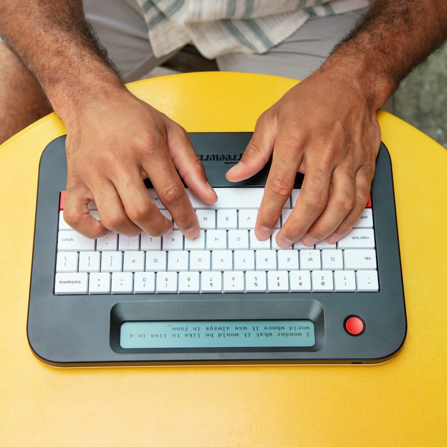 Person typing on a digital typewriter with a screen, next to a mug and books.