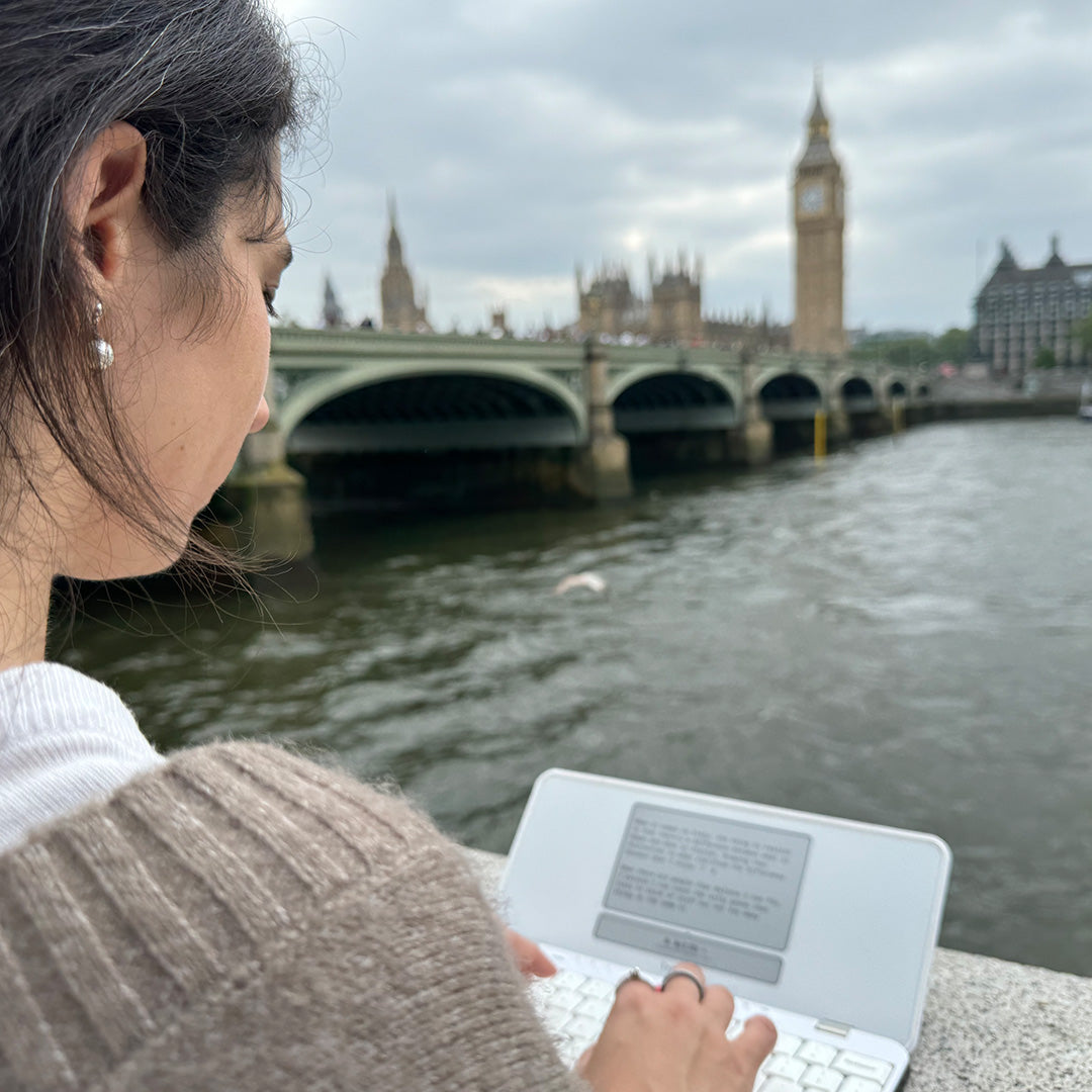 Woman using a laptop by a river with a bridge and a clock tower in the background.